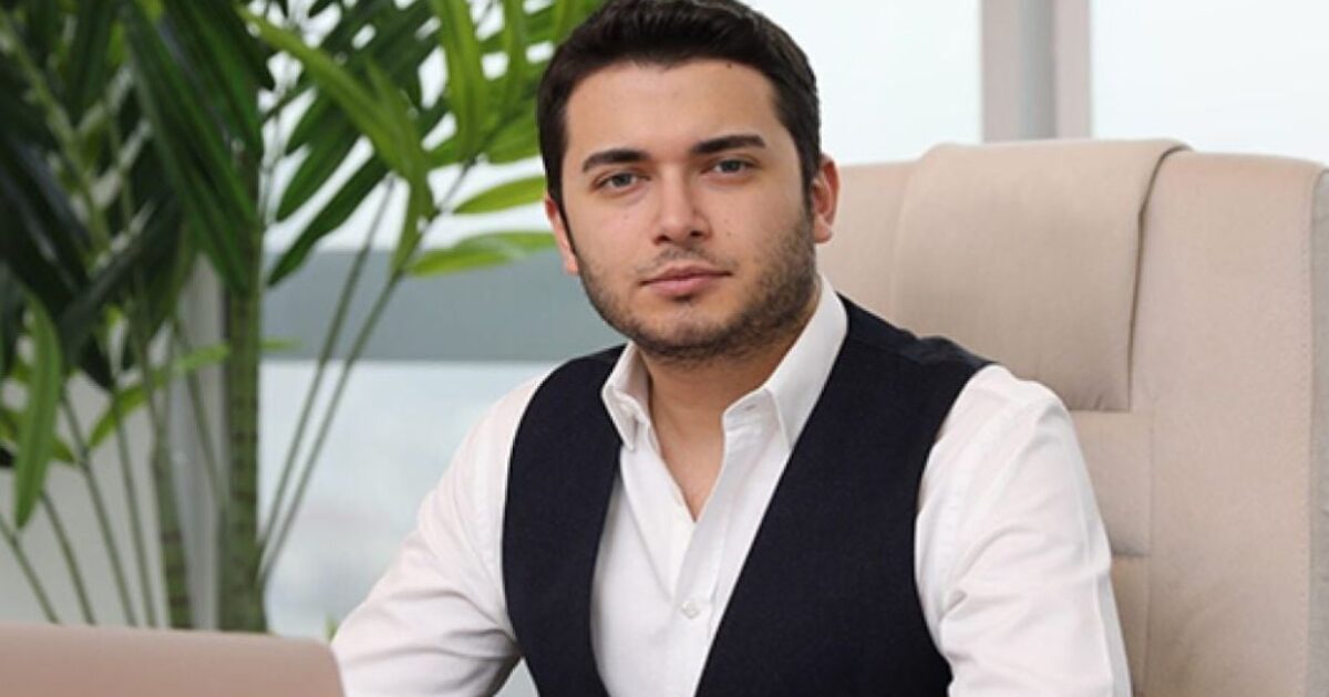 Business professional sitting at a desk with a laptop, surrounded by indoor plants, wearing a white shirt and black vest, conveying a confident demeanor.