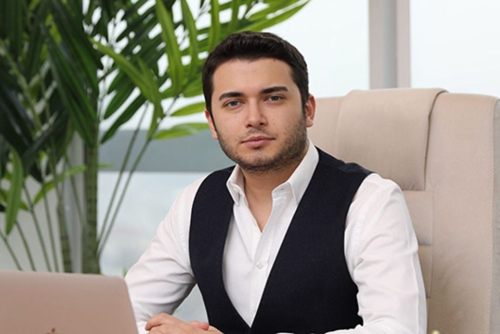 Business professional sitting at a desk with a laptop, surrounded by indoor plants, wearing a white shirt and black vest, conveying a confident demeanor.