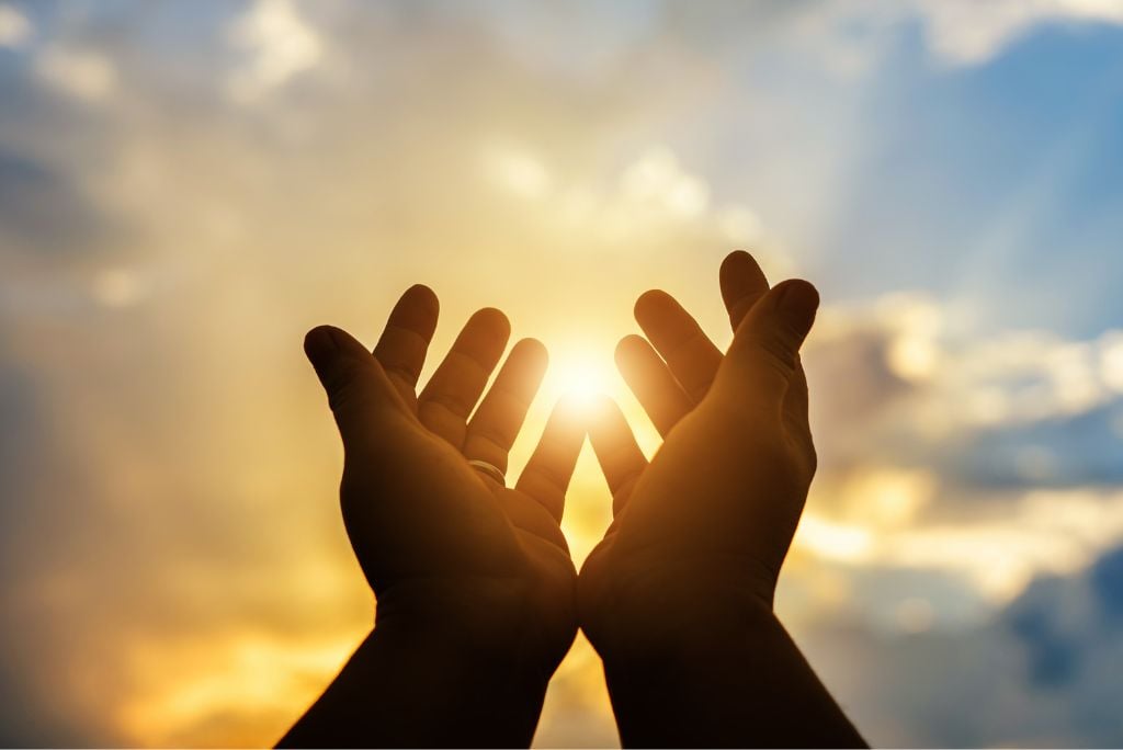 Hands reaching towards a bright sun against a cloudy sky, symbolizing hope and connection with nature.