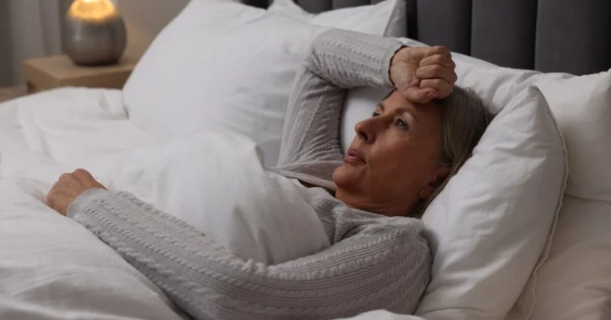 Middle-aged woman lying in bed with a concerned expression, wrapped in blankets, suggesting thoughts of worry or insomnia in a cozy bedroom setting.