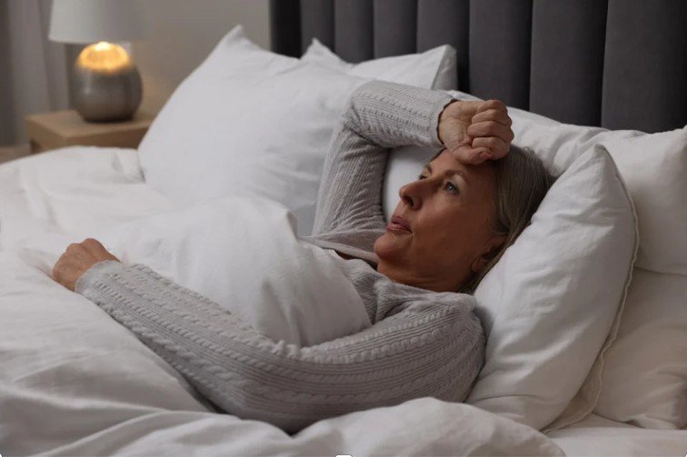 Middle-aged woman lying in bed with a concerned expression, wrapped in blankets, suggesting thoughts of worry or insomnia in a cozy bedroom setting.