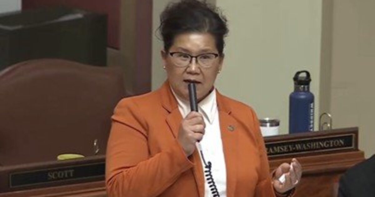 State representative speaking passionately at a legislative session, wearing an orange blazer and holding a microphone, with a water bottle visible in the background.