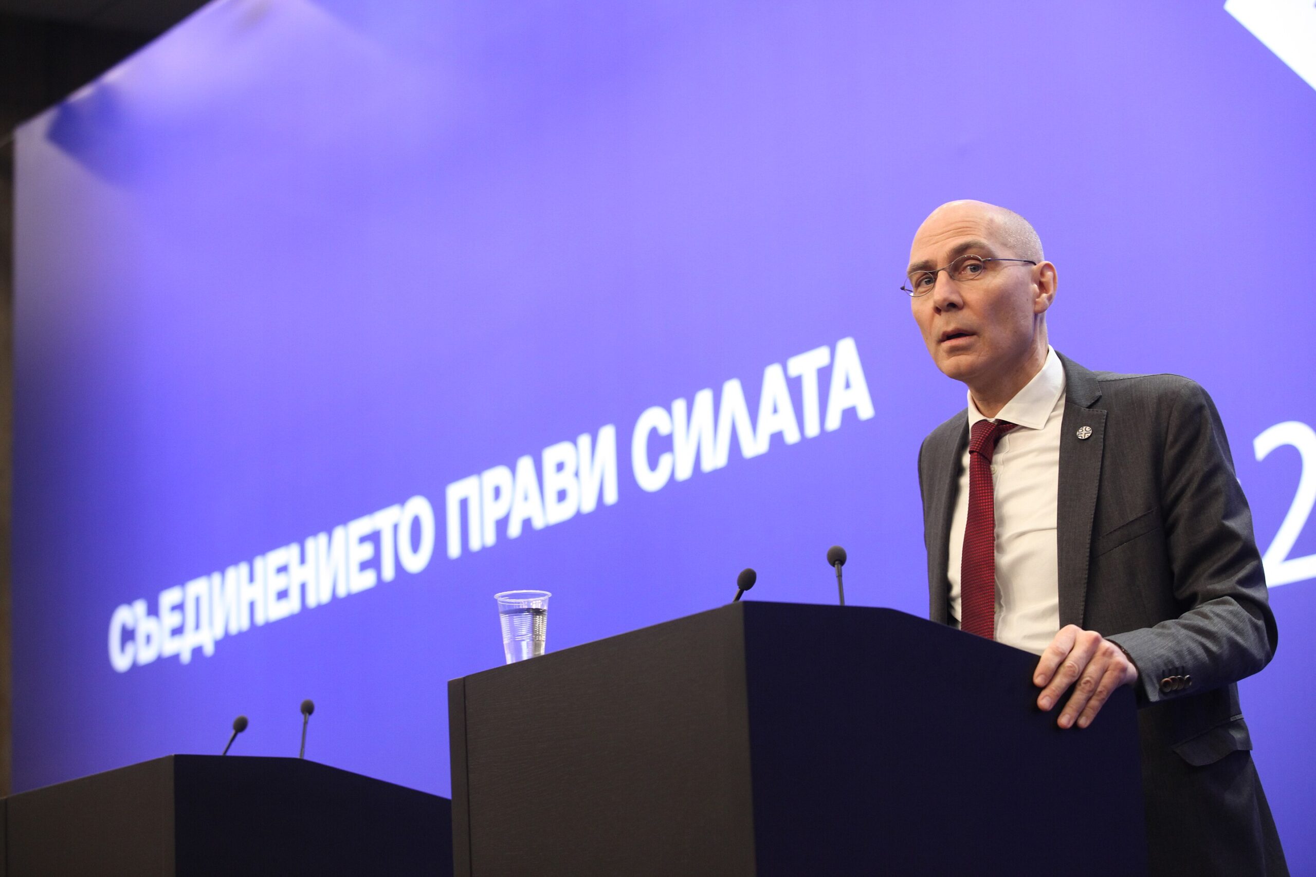 Speaker at a podium addressing an audience, with a backdrop featuring the phrase "Съединението прави силата," emphasizing unity and strength in a conference setting.