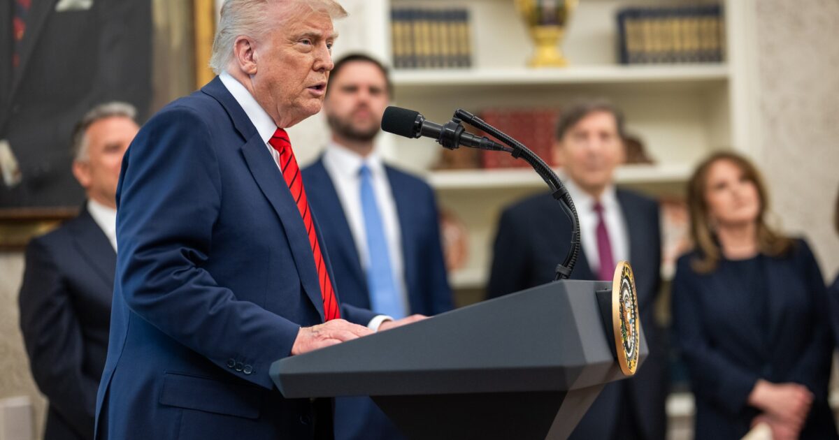 Donald Trump speaks at a podium in the Oval Office, flanked by advisors and a portrait in the background, during a press event.