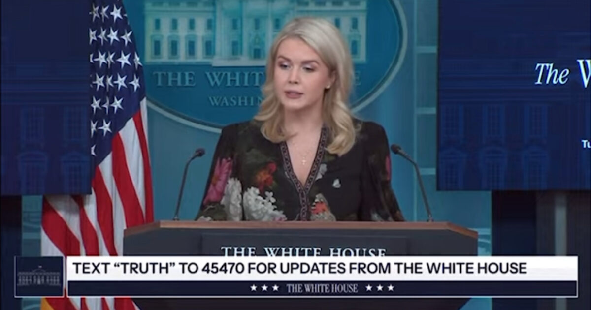 Press briefing at the White House featuring a spokesperson discussing updates, with the American flag displayed prominently in the background.