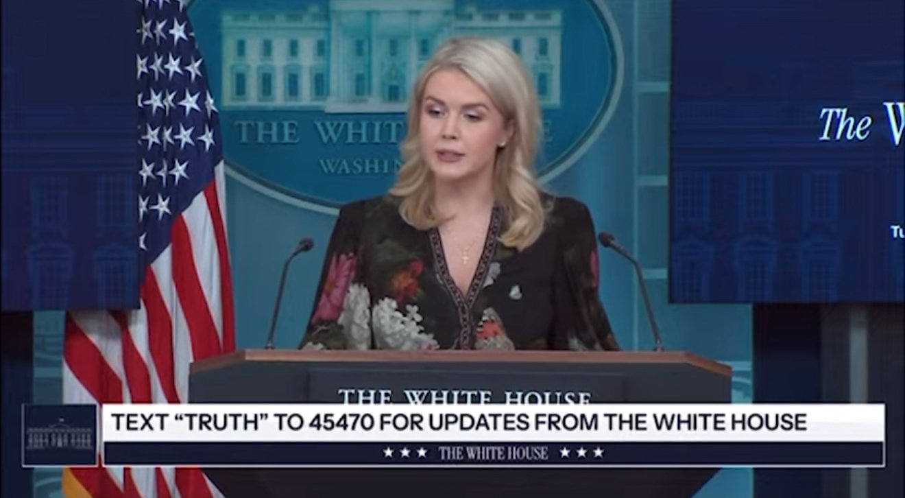 Press briefing at the White House featuring a spokesperson discussing updates, with the American flag displayed prominently in the background.