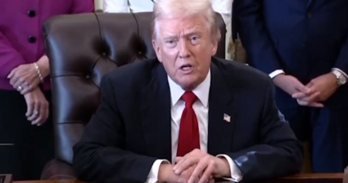 Donald Trump speaking at a table during a formal event with attendees in the background, showcasing a serious expression and wearing a suit and red tie.