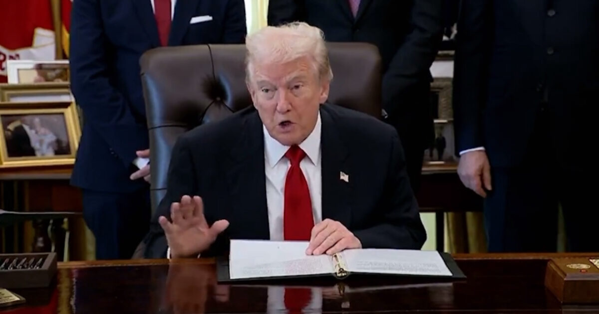 Donald Trump speaking during a formal event in the Oval Office, with officials standing in the background and documents on the desk.
