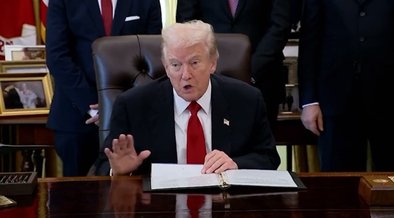 Donald Trump speaking during a formal event in the Oval Office, with officials standing in the background and documents on the desk.