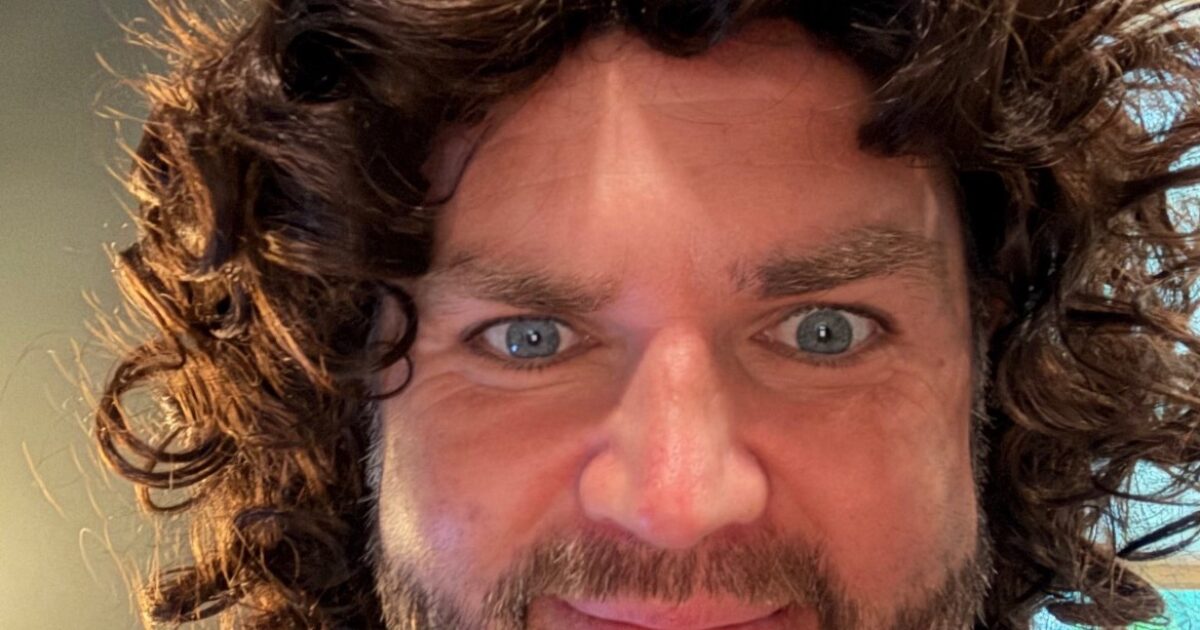Man with curly hair and beard smiling at the camera, showcasing a playful expression and bright blue eyes.