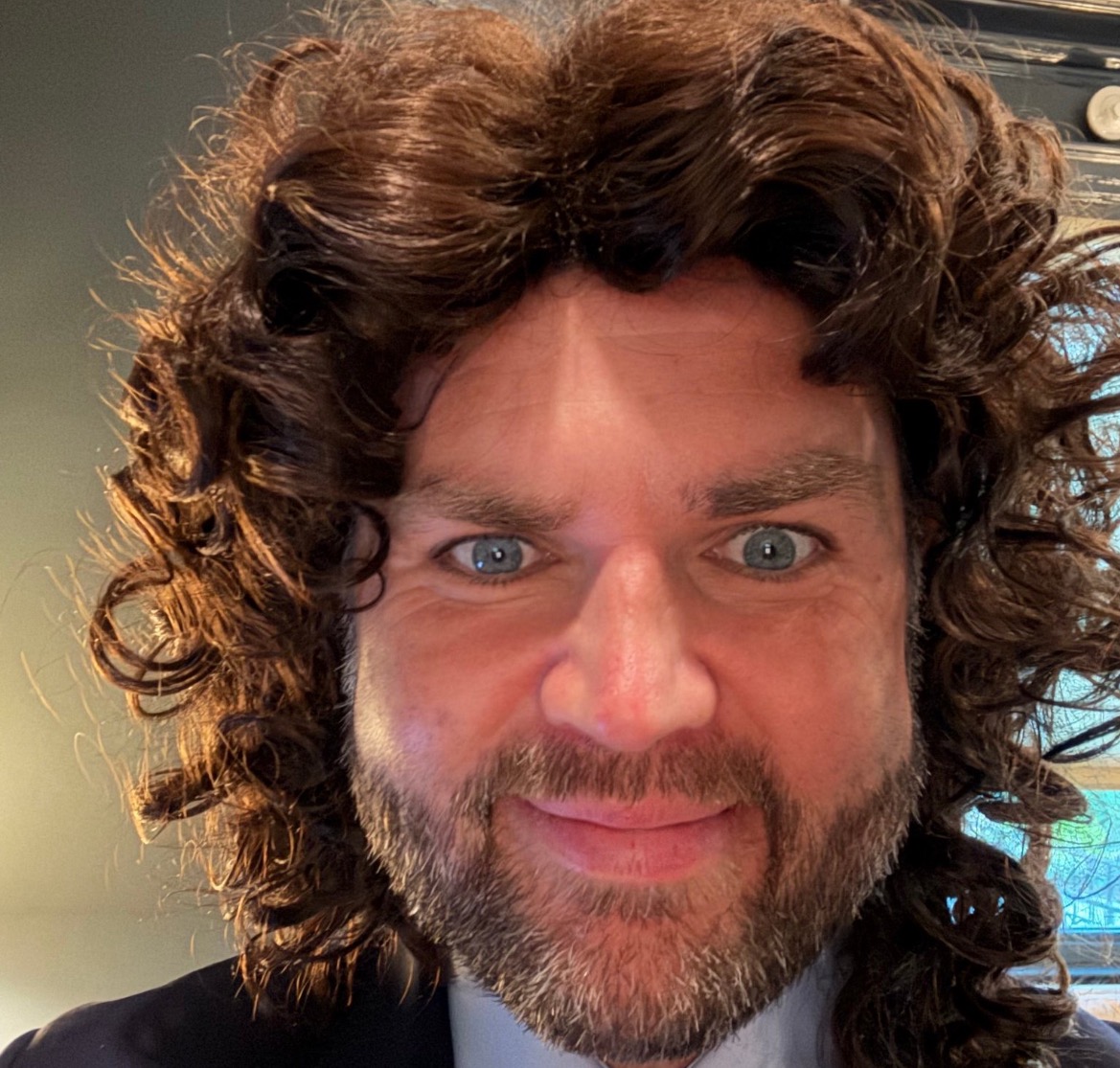 Man with curly hair and beard smiling at the camera, showcasing a playful expression and bright blue eyes.