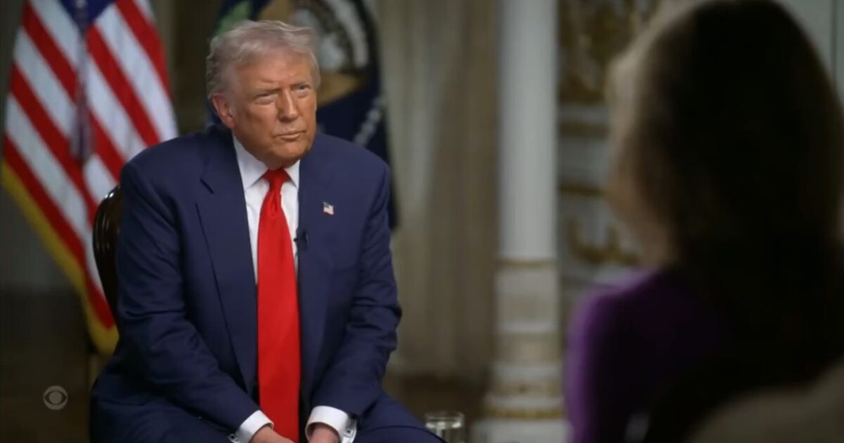 Donald Trump seated for an interview, wearing a blue suit and red tie, with American flags in the background.