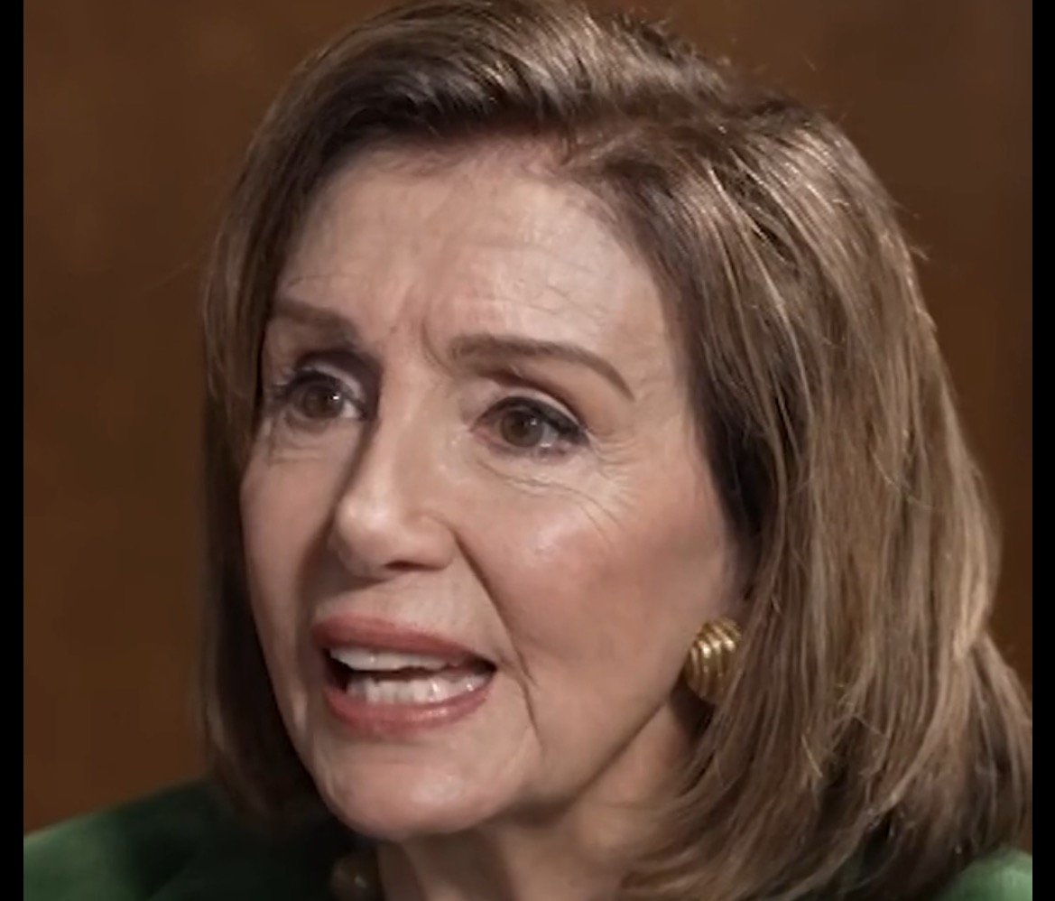 Close-up of a woman with shoulder-length hair, wearing a green outfit and gold earrings, speaking with an expressive demeanor against a neutral background.