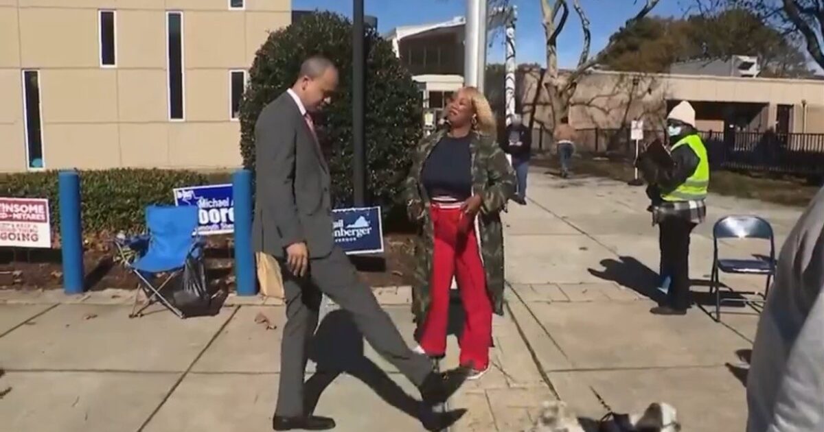 A man and a woman interact playfully outside a polling station, with campaign signs visible in the background during an election event.
