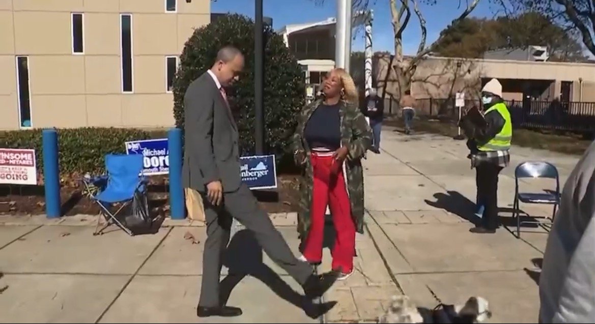 A man and a woman interact playfully outside a polling station, with campaign signs visible in the background during an election event.