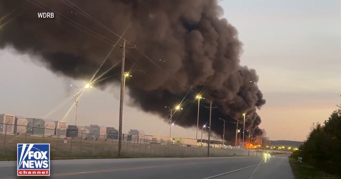 Thick black smoke billows from a fire near industrial buildings along a road, highlighting a significant fire incident in the area.