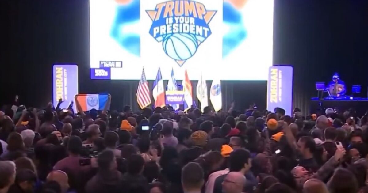 Crowd gathered at a political event featuring a large "Trump is Your President" banner, with flags and a DJ in the background.