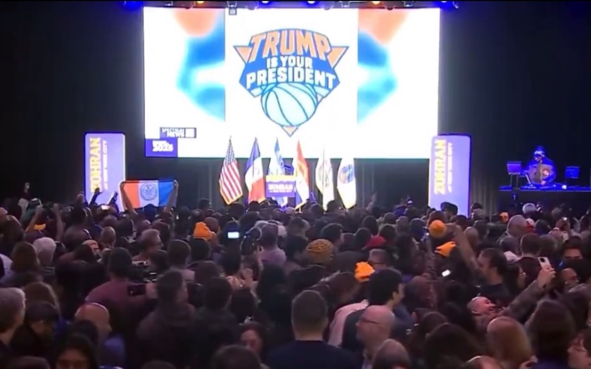 Crowd gathered at a political event featuring a large "Trump is Your President" banner, with flags and a DJ in the background.