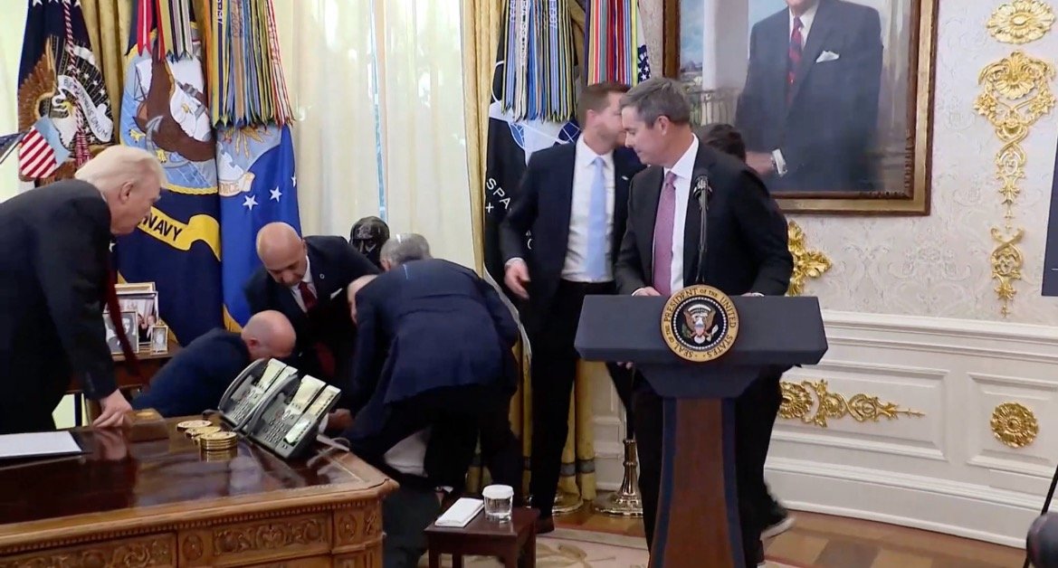 A group of officials gathers around a table in the Oval Office, focusing on a situation involving a seated individual.