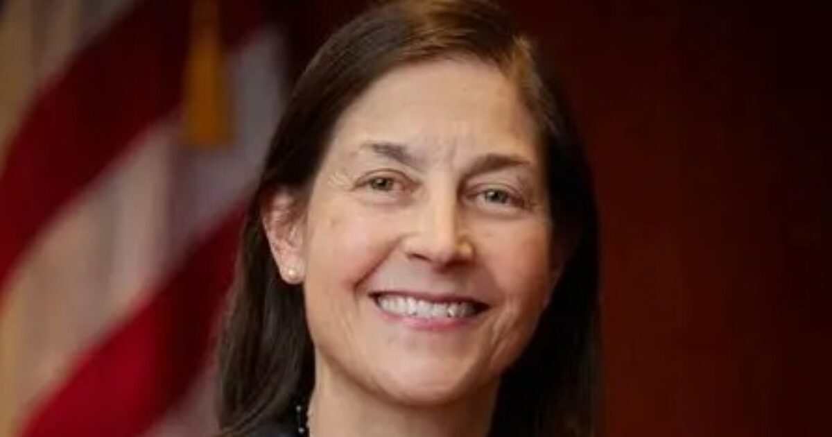 Smiling woman with dark hair and earrings, standing in front of an American flag backdrop, exuding confidence and professionalism.
