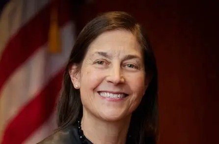 Smiling woman with dark hair and earrings, standing in front of an American flag backdrop, exuding confidence and professionalism.