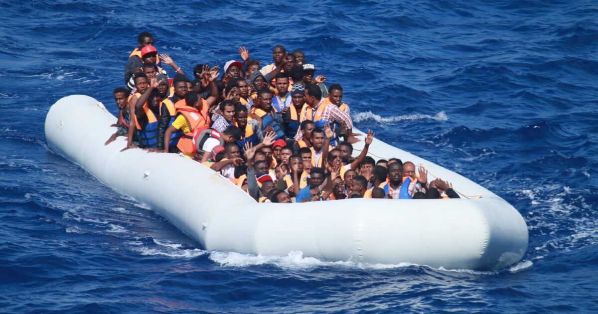 Group of migrants in life jackets crowded on an inflatable boat in the ocean, signaling for help amidst a humanitarian crisis.