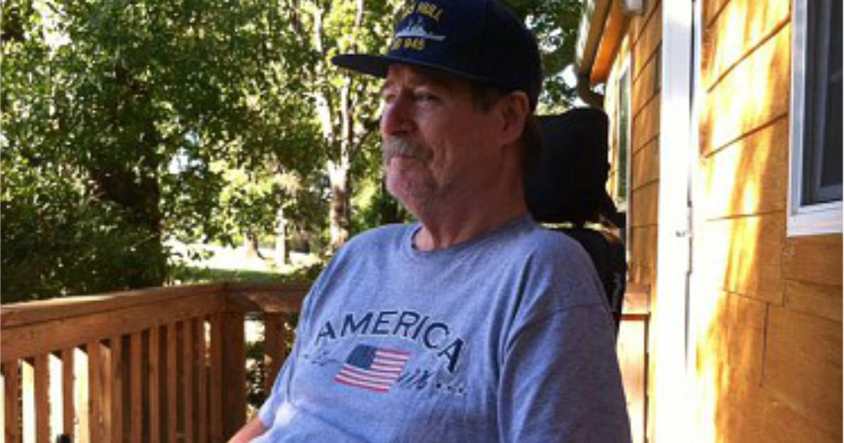 A man in a wheelchair wearing a gray "America" t-shirt and a cap sits on a wooden porch surrounded by greenery.