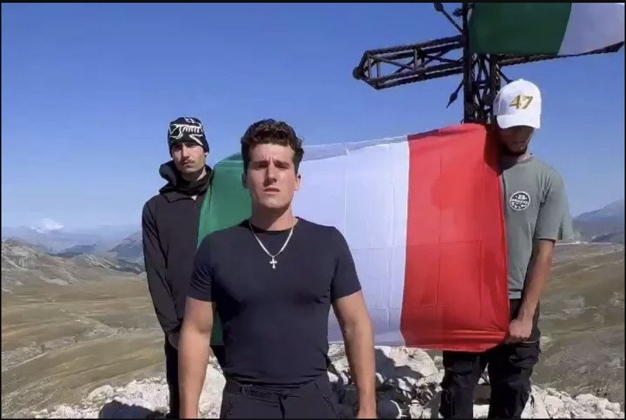 Three young men pose on a mountain peak, proudly displaying the Italian flag against a scenic backdrop.