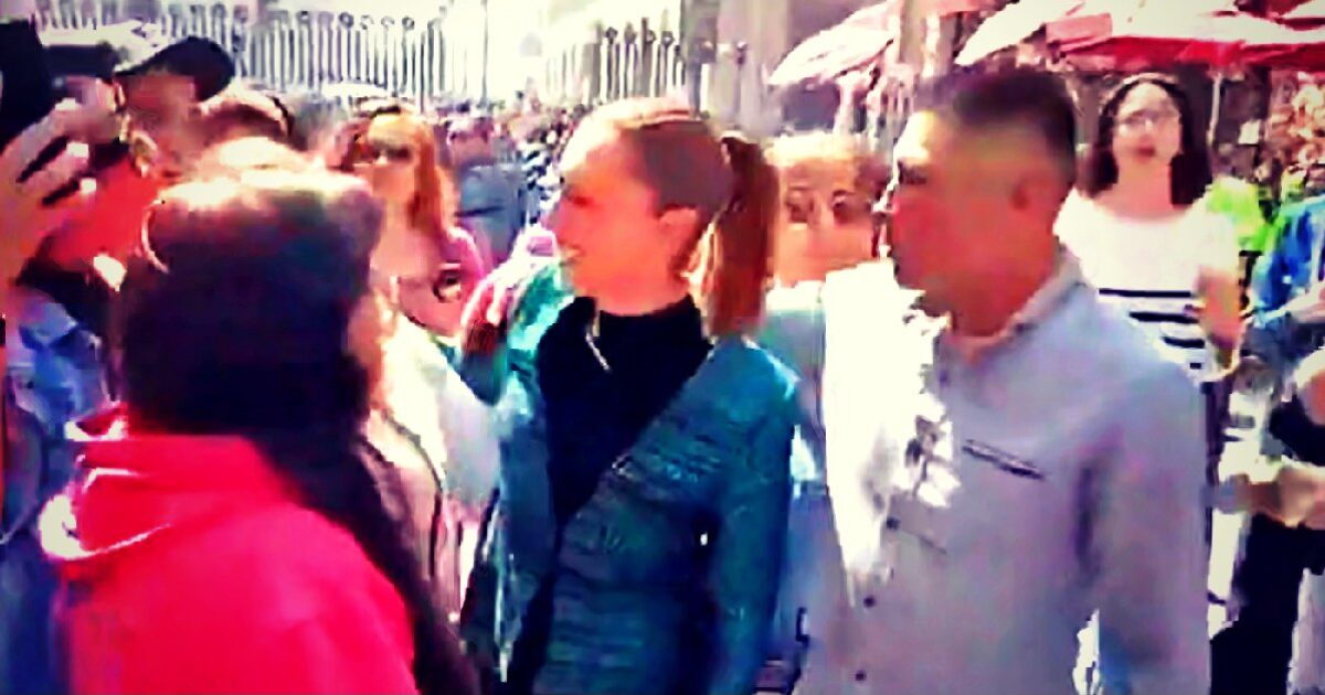 A crowd of people interacting in a vibrant outdoor setting, with two individuals at the forefront engaged in conversation amidst the lively atmosphere.