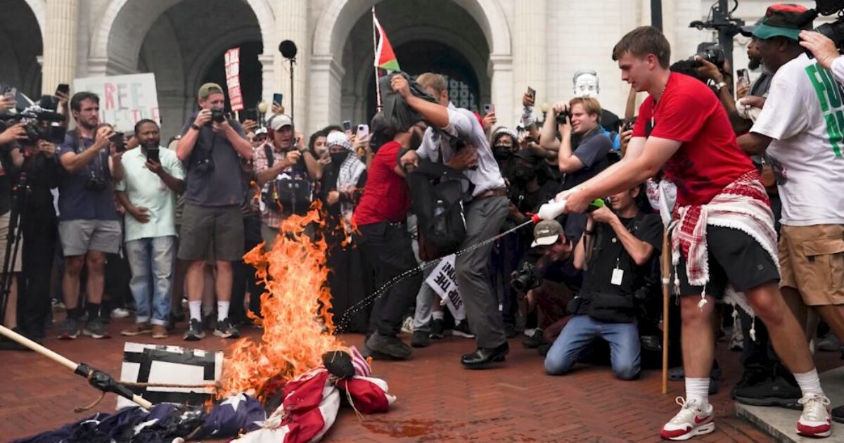 Protesters gather as a person sets fire to a flag during a demonstration, surrounded by media and onlookers capturing the event.