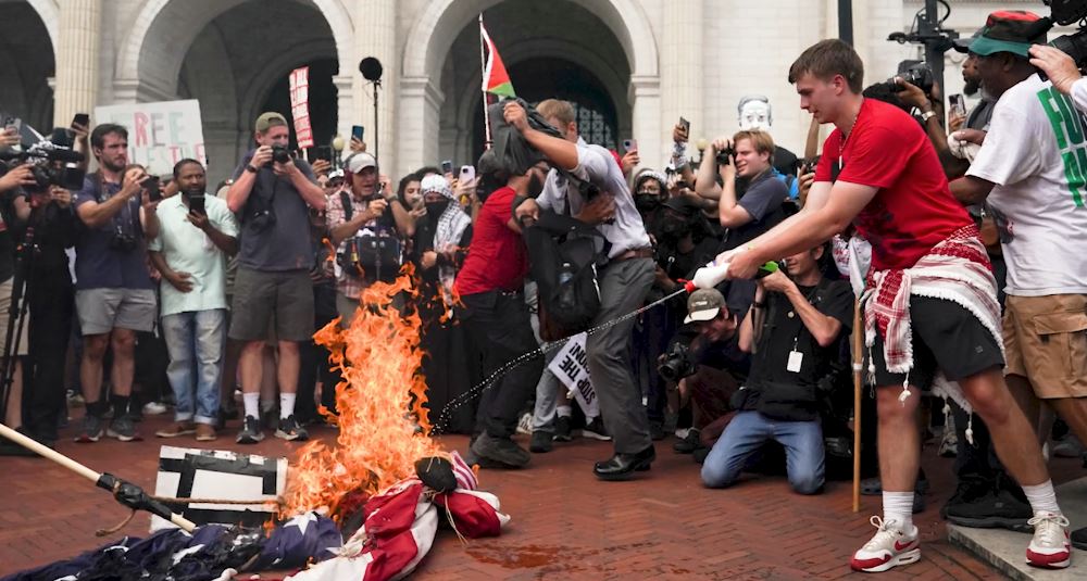 Protesters gather as a person sets fire to a flag during a demonstration, surrounded by media and onlookers capturing the event.
