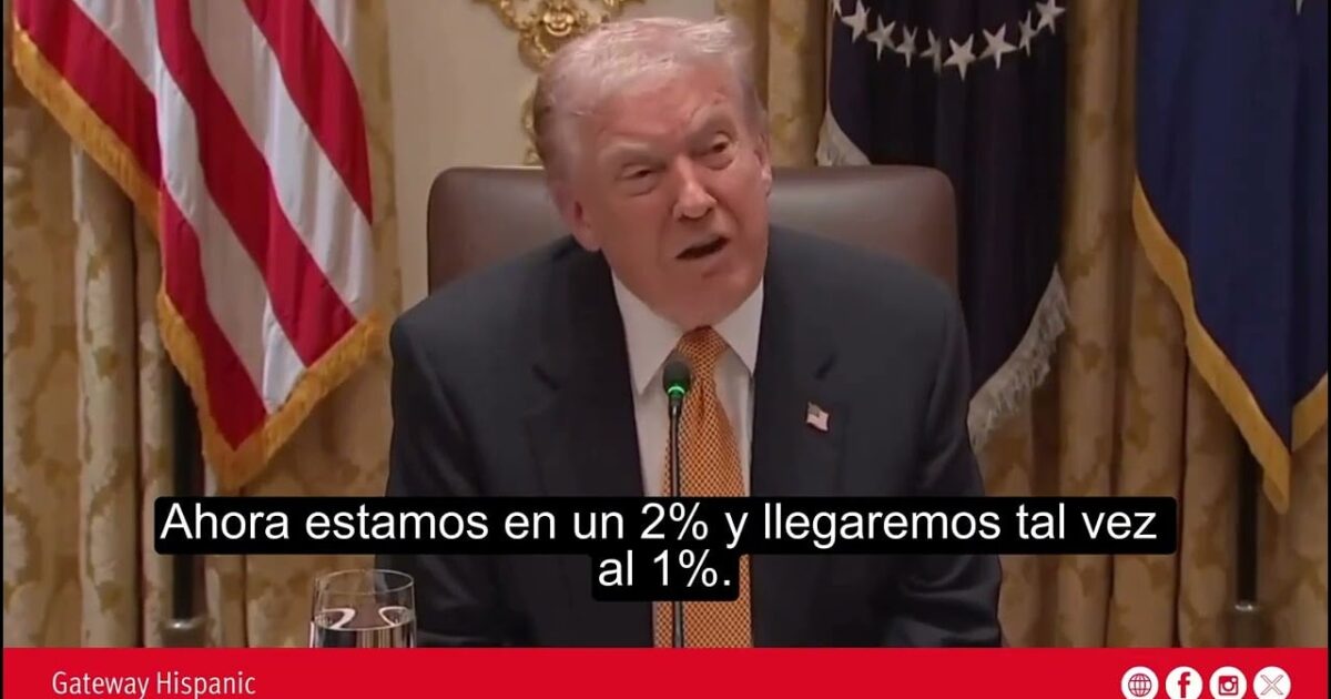 Donald Trump speaking during a meeting, discussing economic percentages, with U.S. flags in the background.