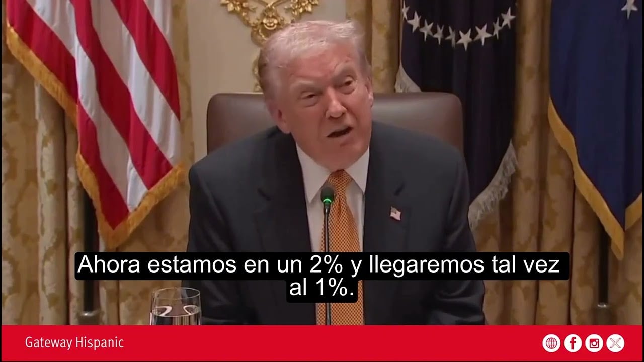 Donald Trump speaking during a meeting, discussing economic percentages, with U.S. flags in the background.