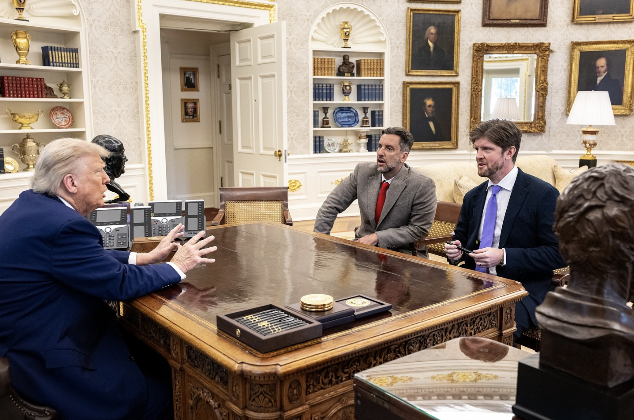 Donald Trump engages in a discussion with two men at the Resolute Desk in the Oval Office, surrounded by historical portraits and office decor.