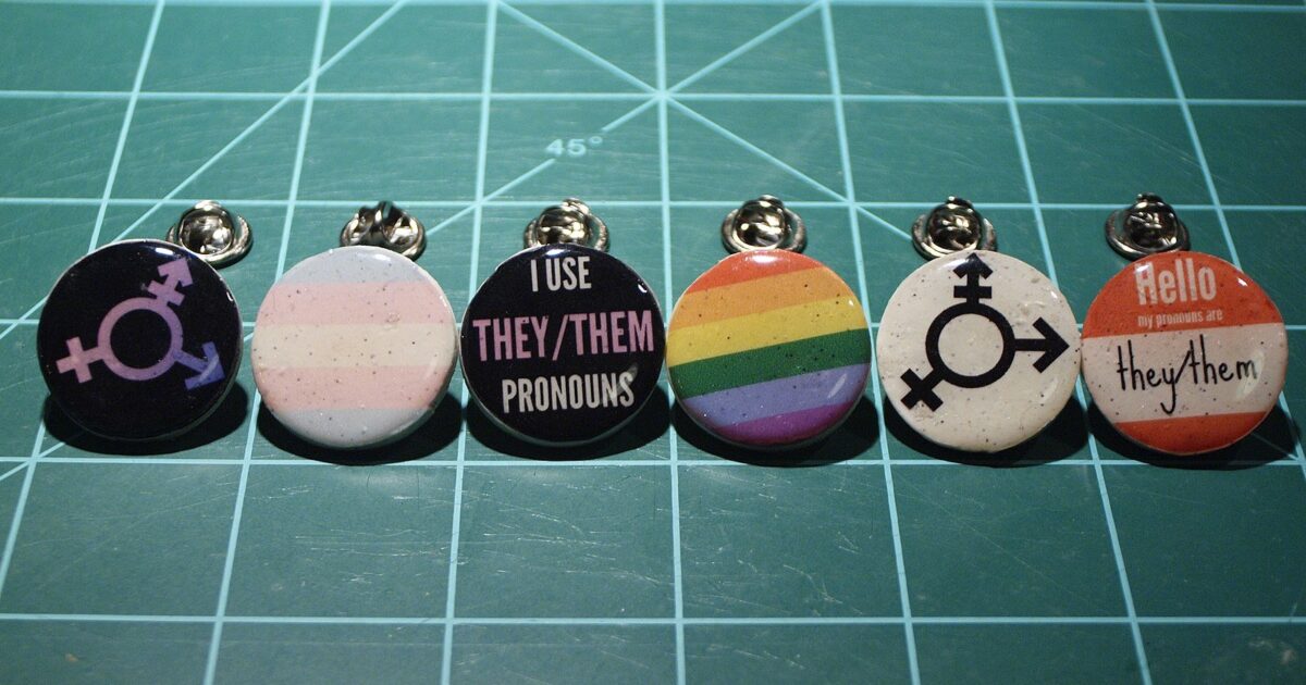 Colorful pins featuring gender symbols and pronouns, promoting inclusivity and awareness of they/them pronouns on a green cutting mat.