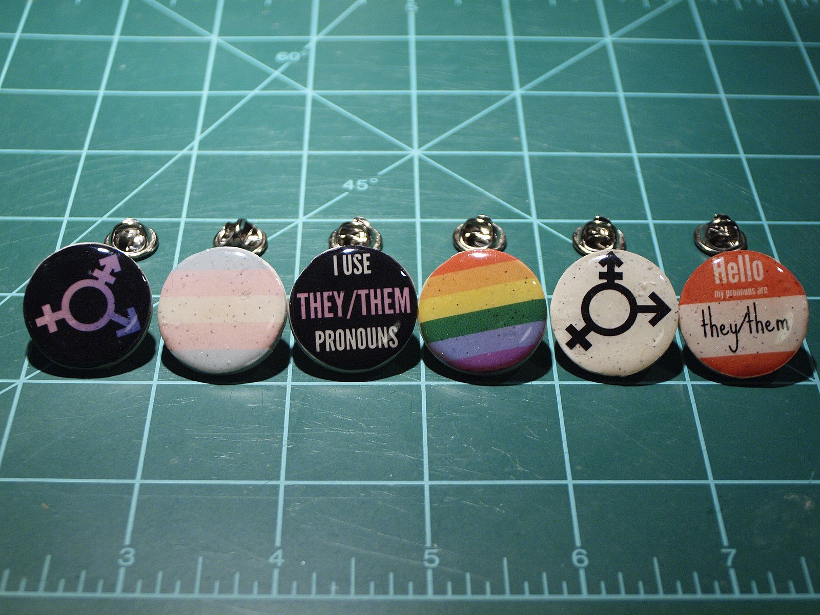 Colorful pins featuring gender symbols and pronouns, promoting inclusivity and awareness of they/them pronouns on a green cutting mat.