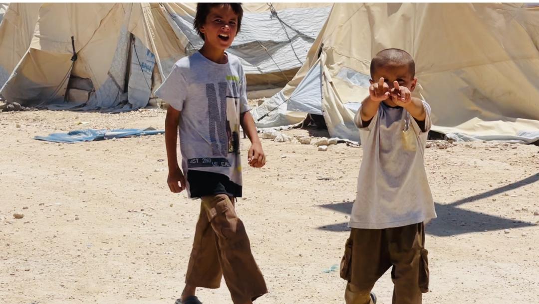 Two young boys play and interact in a refugee camp, surrounded by tents and a sandy environment.