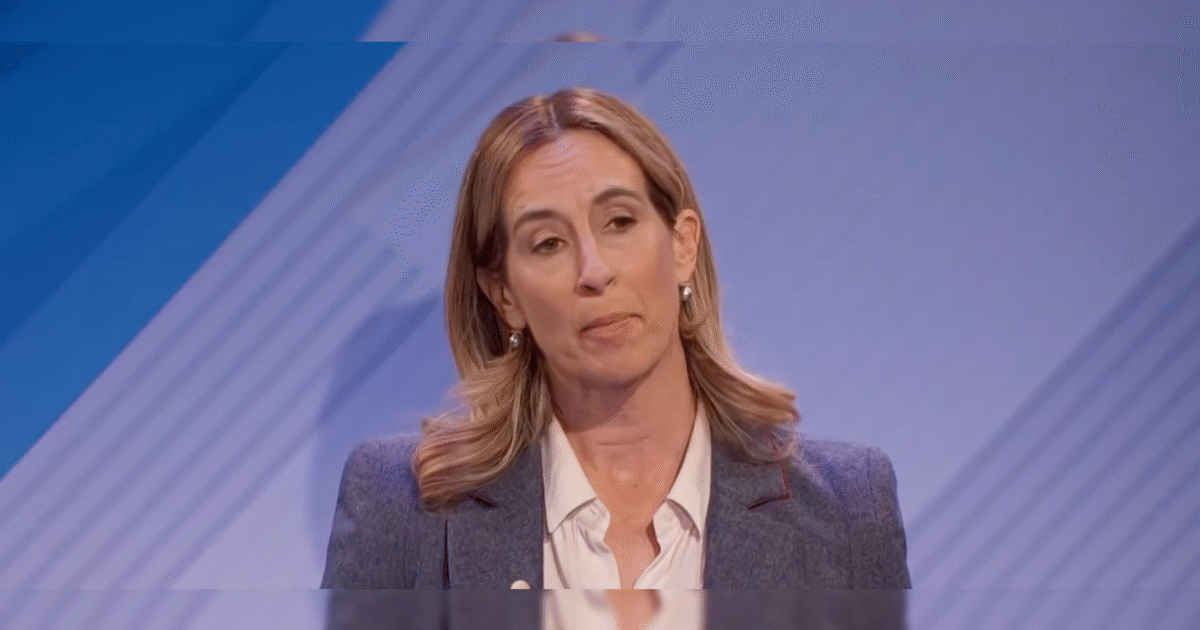 Woman with shoulder-length hair speaking during a presentation, wearing a gray blazer and white shirt against a blue background.