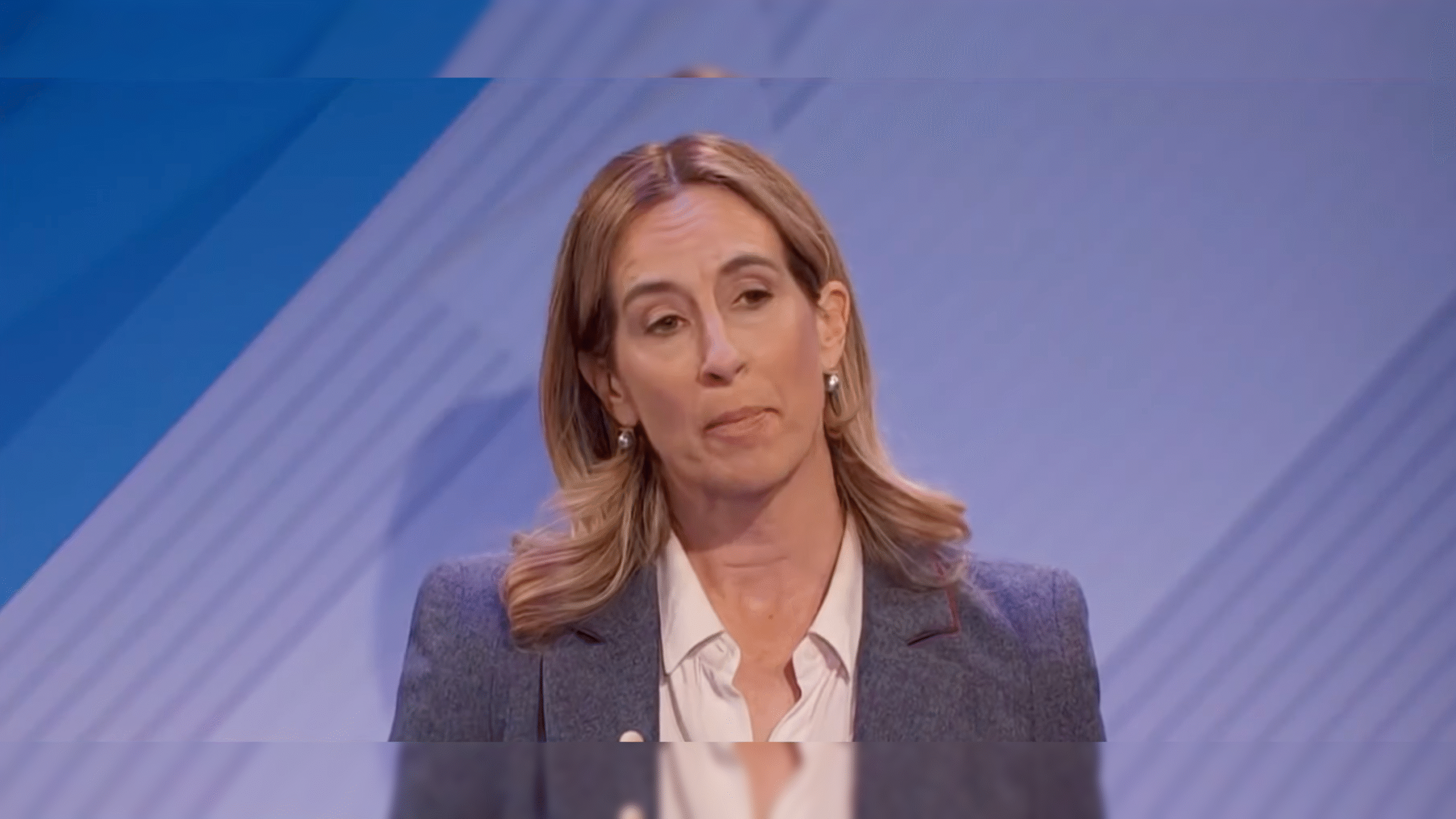 Woman with shoulder-length hair speaking during a presentation, wearing a gray blazer and white shirt against a blue background.