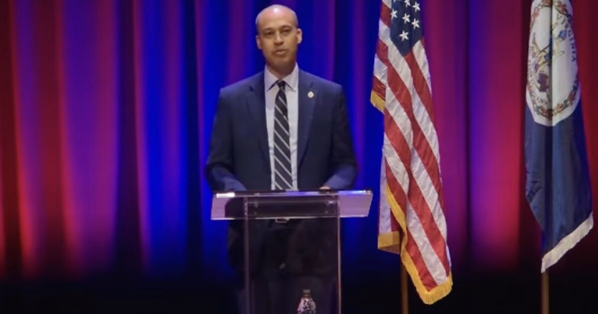 A speaker stands at a podium with American and Virginia flags in a well-lit auditorium, addressing an audience during a formal event.