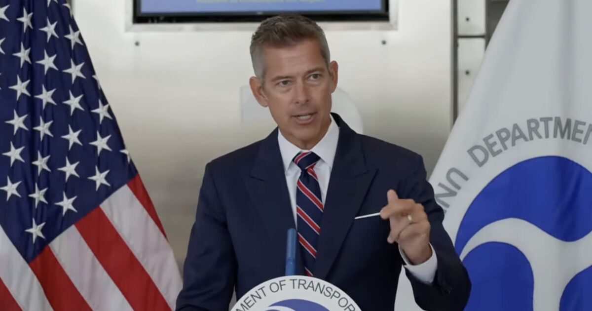 Man in a suit speaking at a podium with the Department of Transportation logo, flanked by an American flag, during a press event.