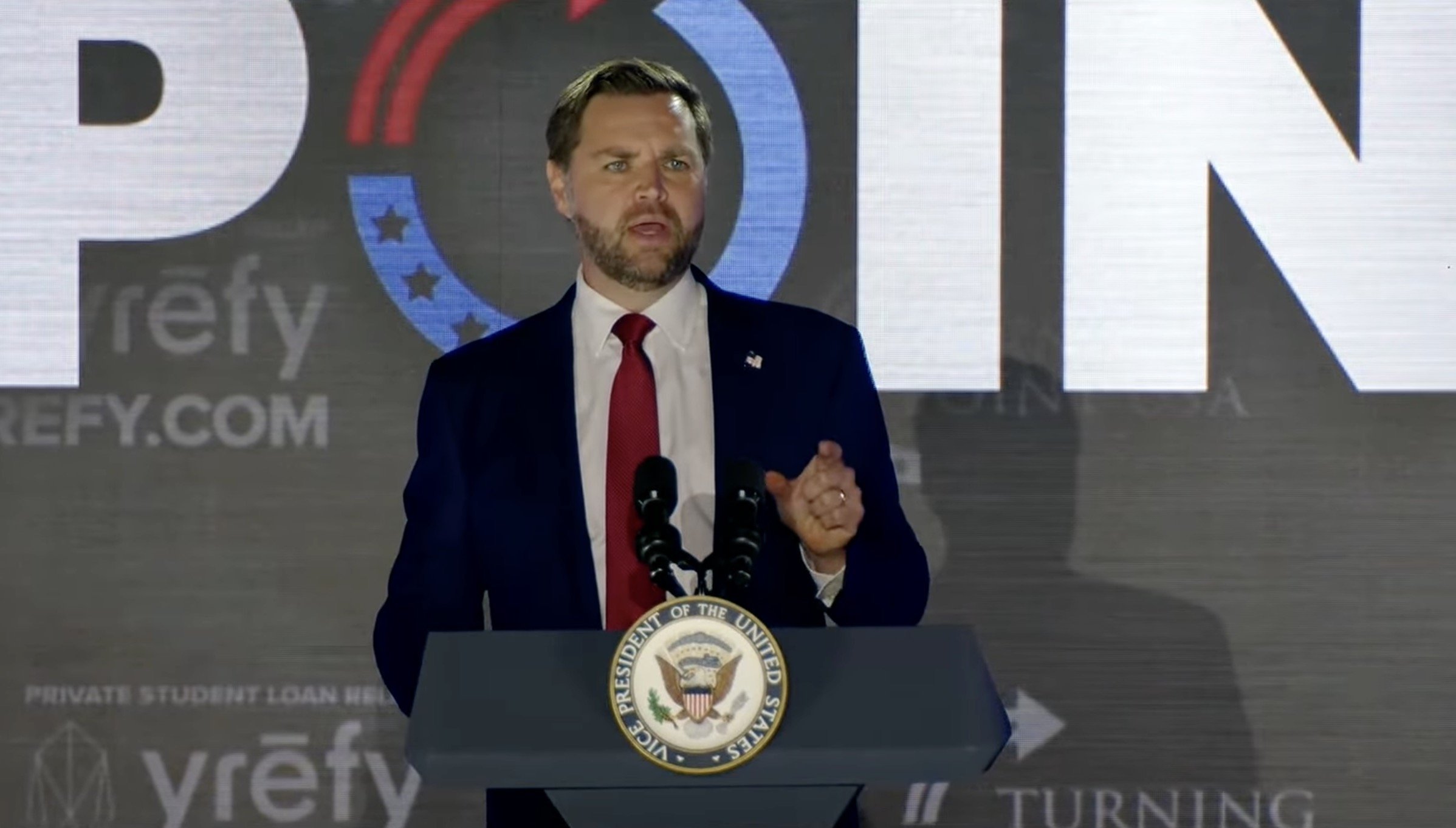Speaker at a podium with the Vice President seal, addressing an audience during a political event, with a large backdrop displaying the word "POINT."