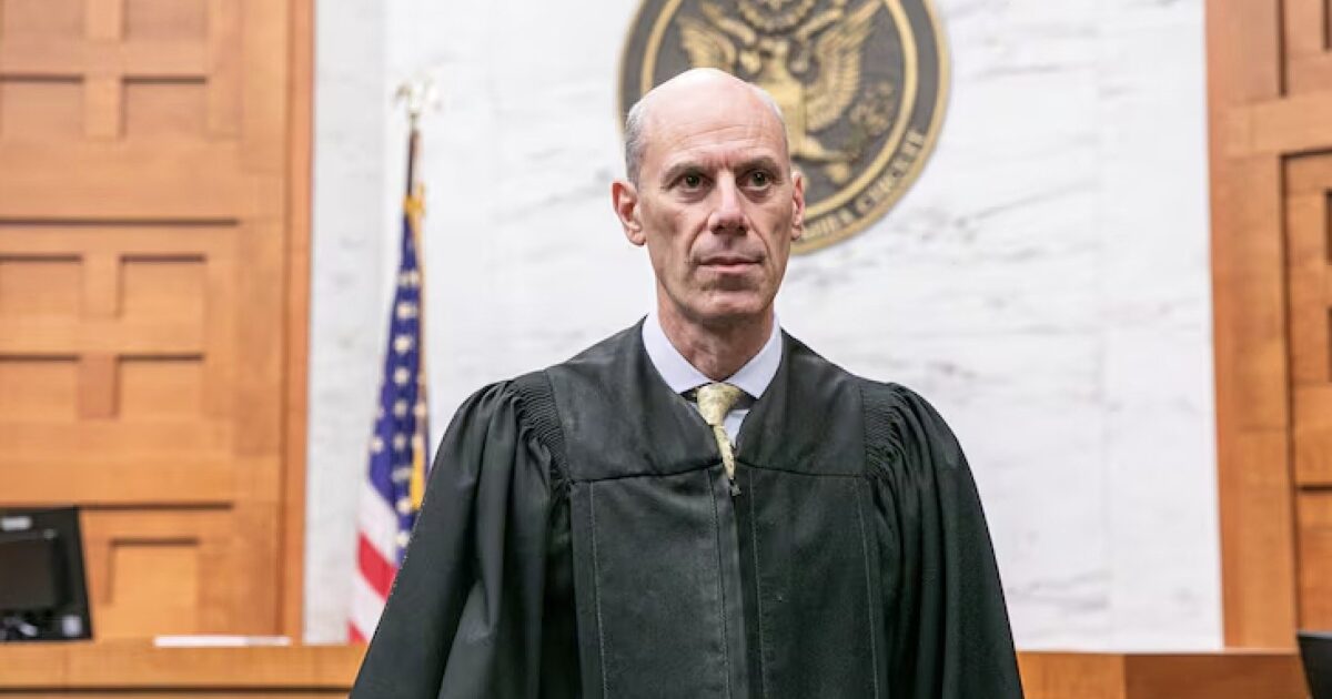 Portrait of a judge in a courtroom, wearing a black robe, with an American flag and court seal in the background.