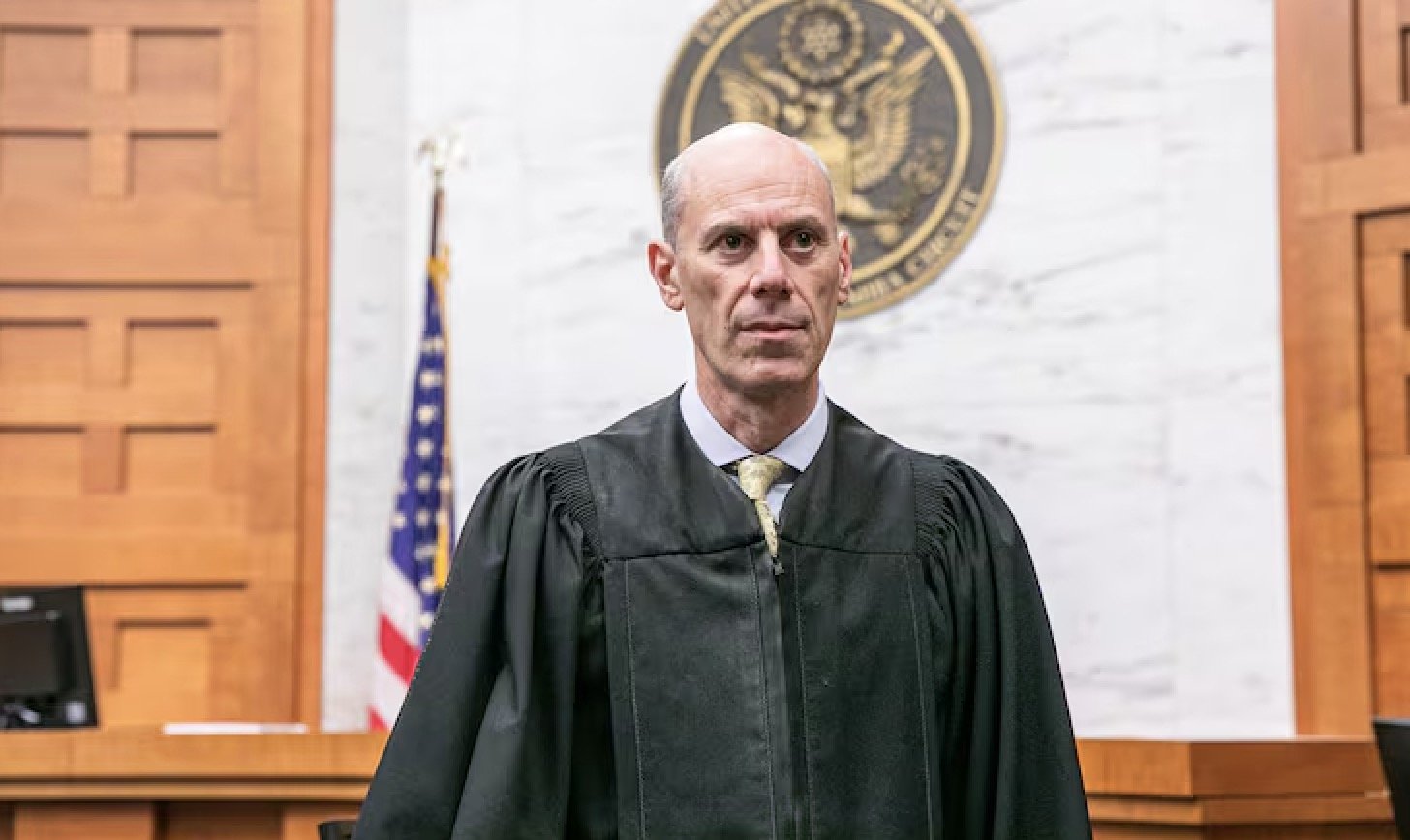 Portrait of a judge in a courtroom, wearing a black robe, with an American flag and court seal in the background.