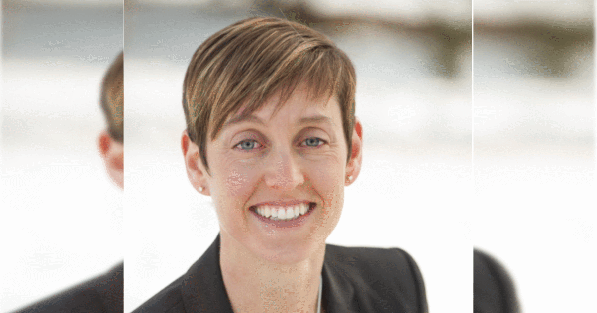 Smiling woman with short hair wearing a black blazer, standing outdoors against a blurred background, exuding confidence and approachability.