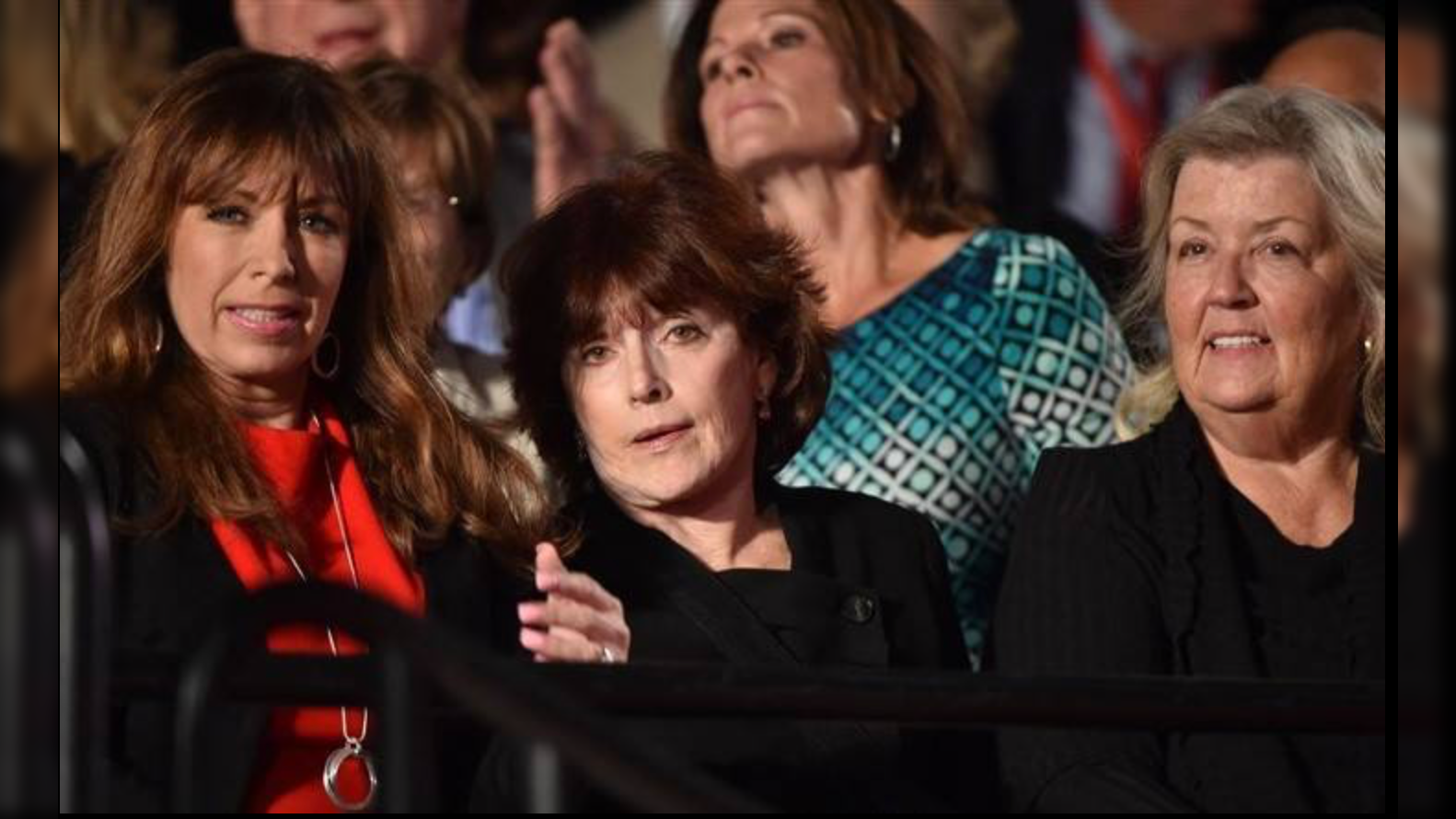 Three women seated in an audience, engaged in conversation, with a crowd visible in the background during a live event.