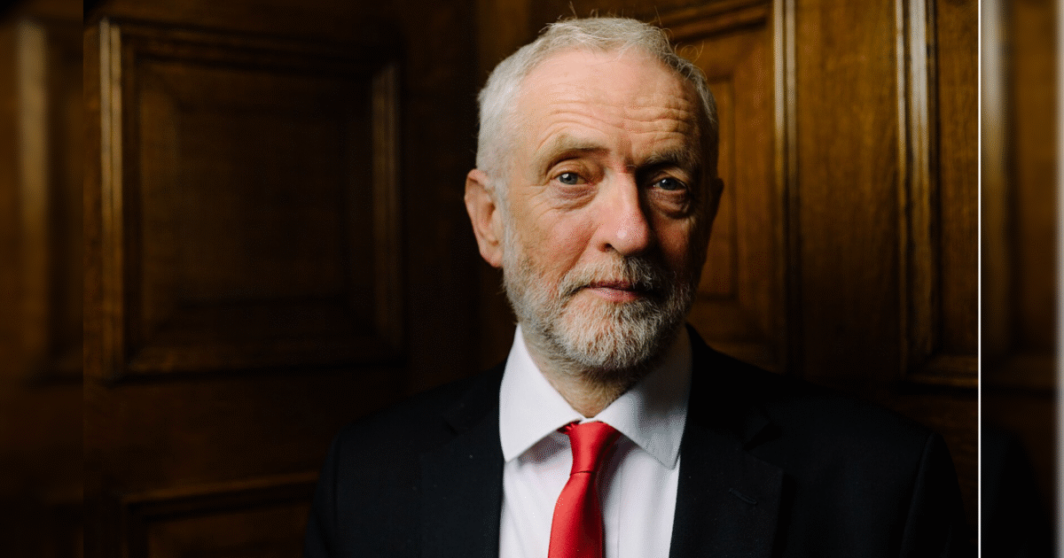 Portrait of a man with a beard and gray hair, wearing a black suit and red tie, standing against a wooden background.