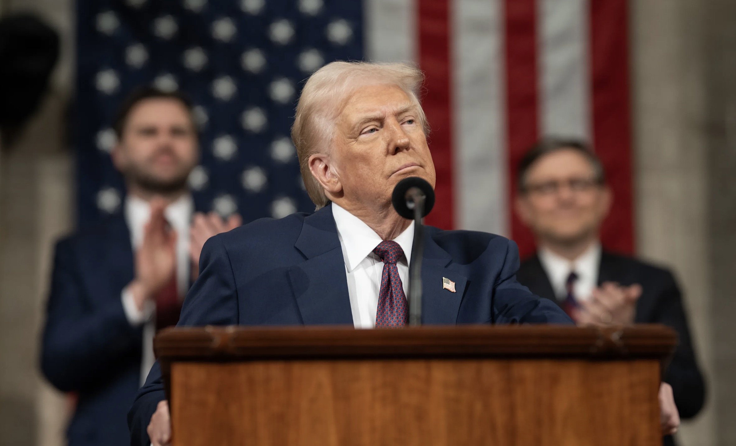 Donald Trump delivers a speech at a podium with the American flag in the background, while supporters applaud in the audience.