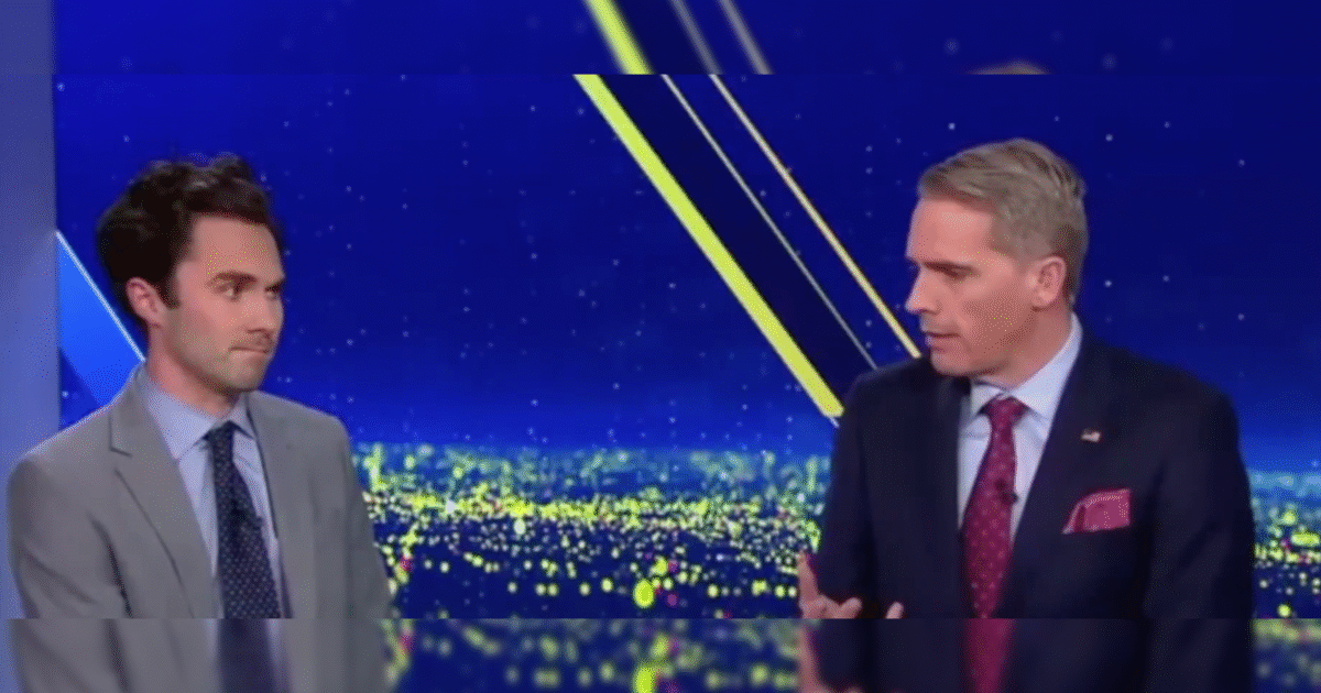 Two male news anchors engage in a discussion on a brightly lit television set with a cityscape background.