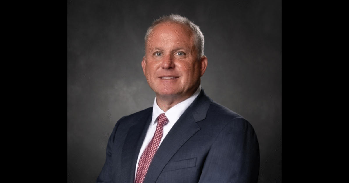Professional headshot of a smiling man in a dark suit and tie, set against a neutral gray background.